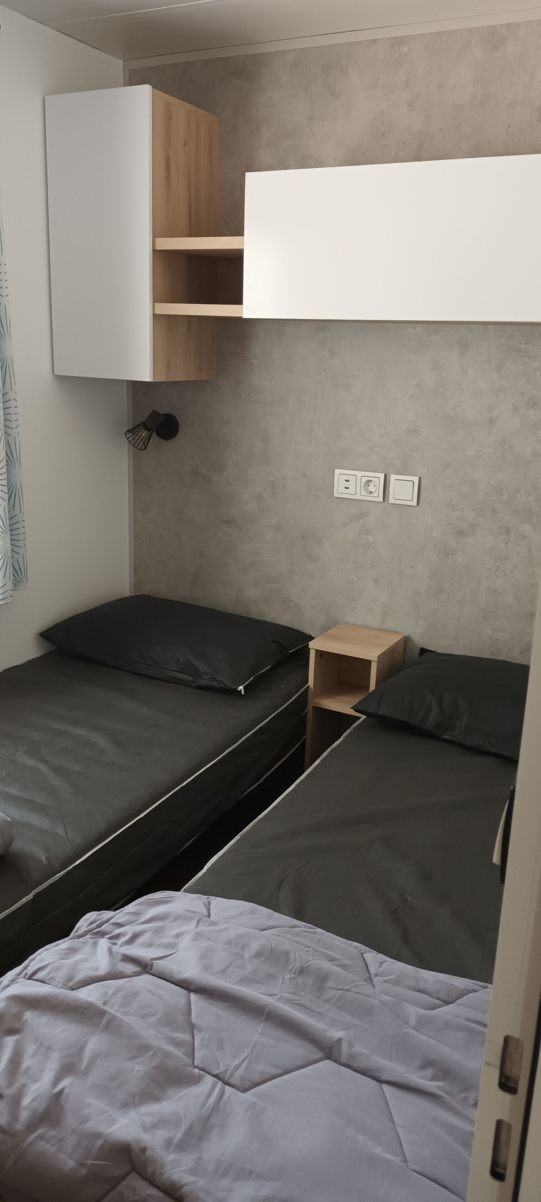 Una habitación compacta con dos camas, una mesita de noche de madera y un armario fijo en la pared.