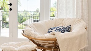 A wicker chair with a cushion, a patterned pillow, and a white blanket, positioned near a window with a view of a fence and greenery. - Luxury Villa | Casa WED | Bahia Beach PR (Rio Grande)