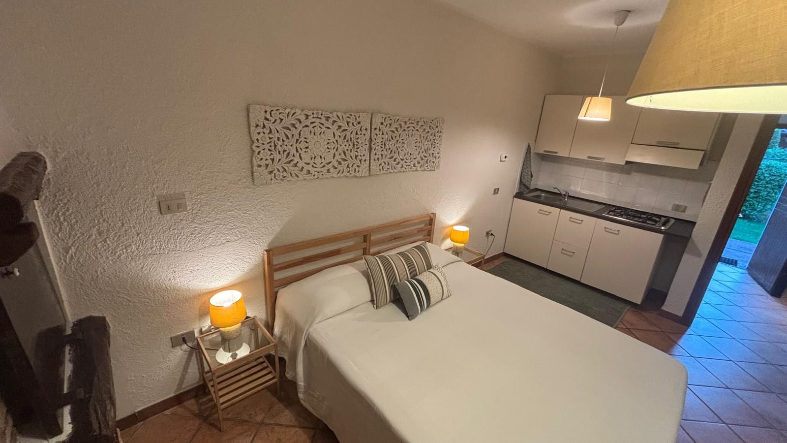 A bedroom with a bed, bedside lamps, a nightstand, and a kitchenette in the background.