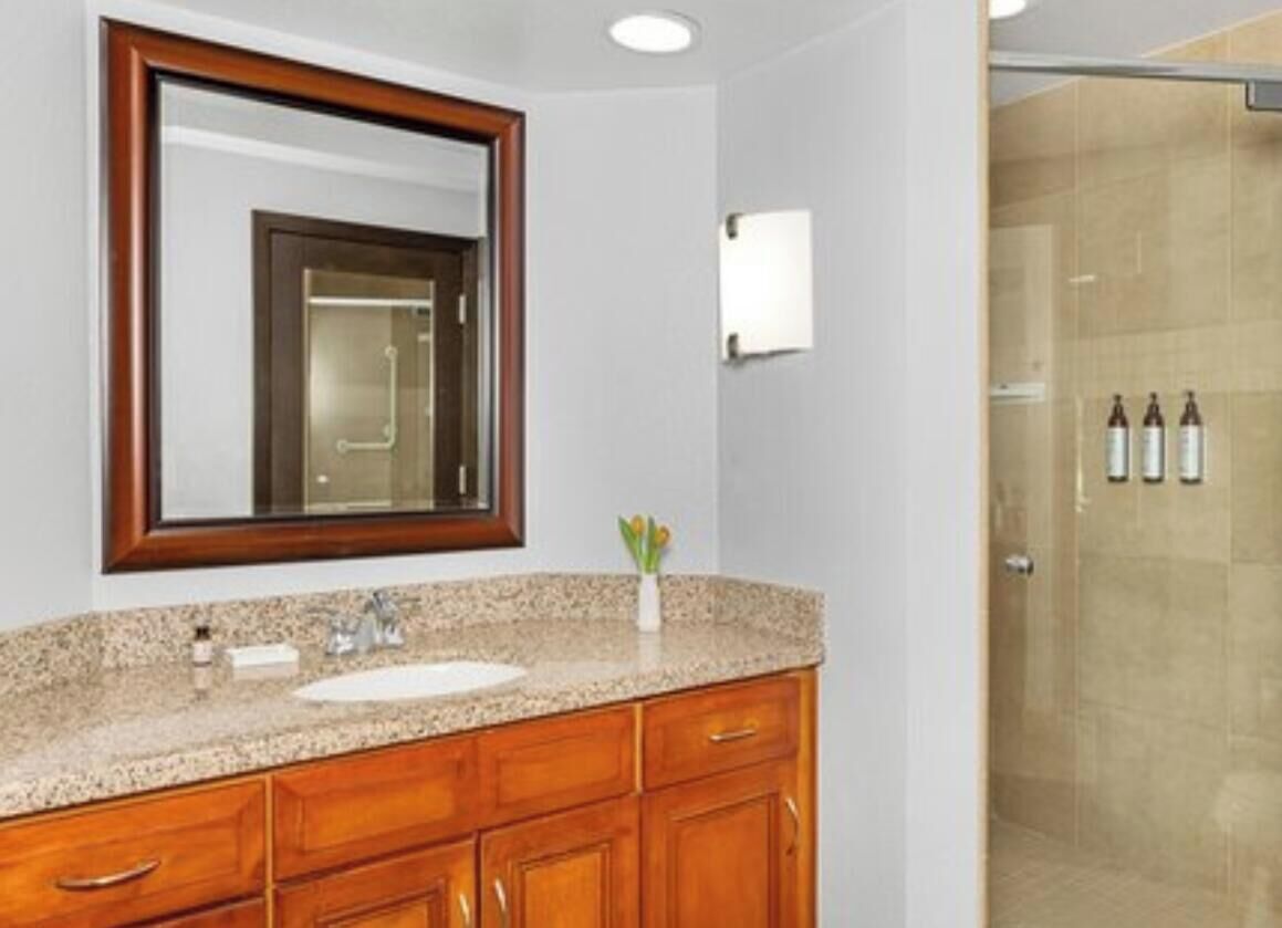 A bathroom with a granite countertop, wooden cabinets, a mirror, a sink, and a glass shower enclosure.