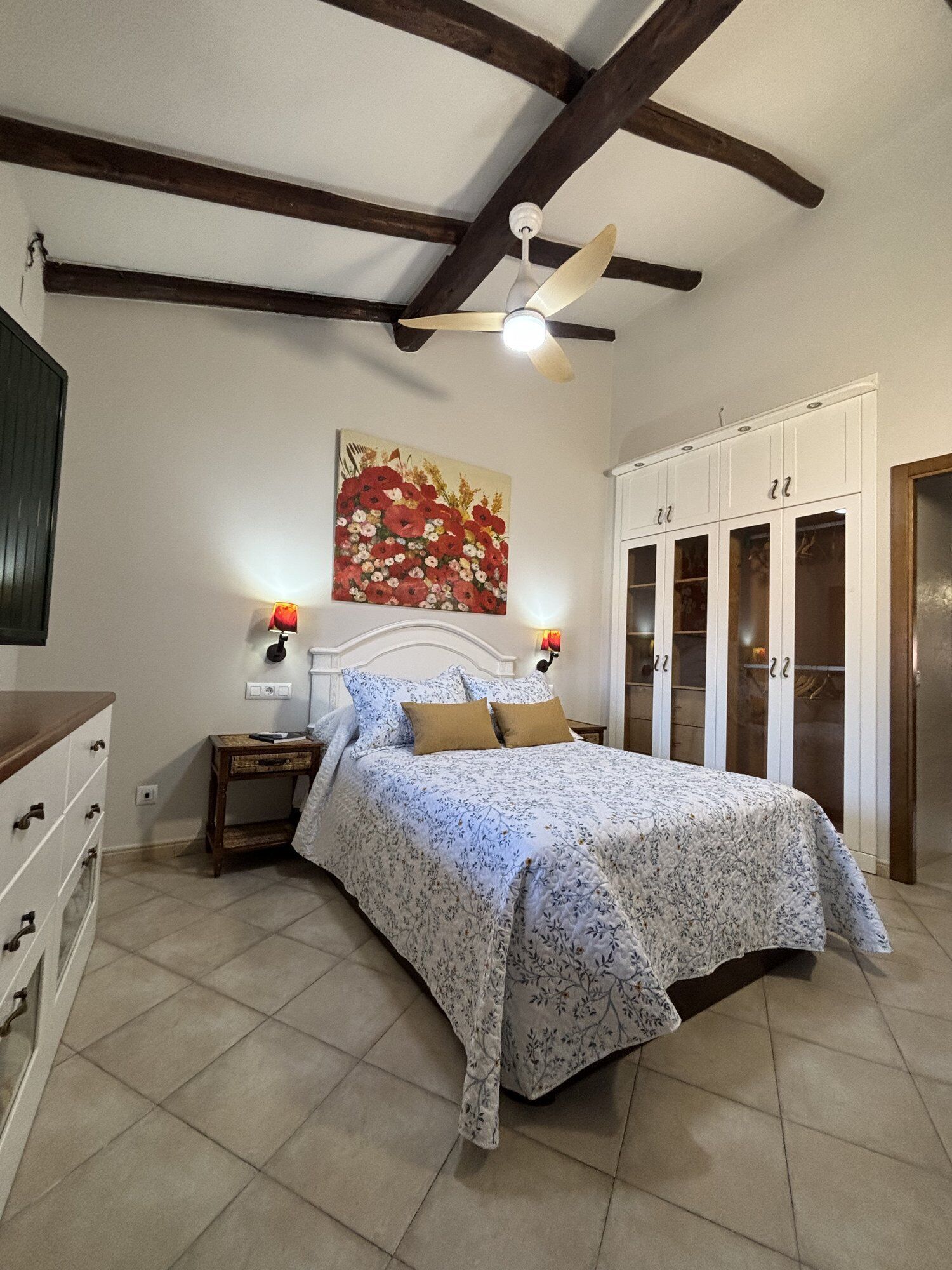 A bedroom with a bed, a nightstand, a ceiling fan, a wardrobe, and a painting on the wall.