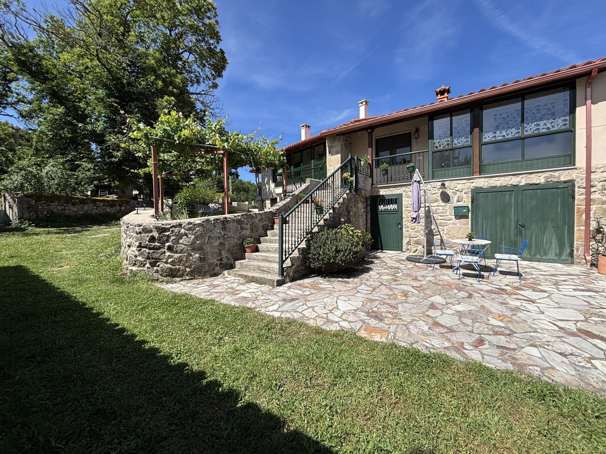 A stone house with a stone staircase leading to a green door, a small garden with grass and trees, and a clear blue sky.