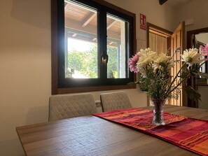 A dining area with a table set for two, a vase of flowers on the table, and a window with a view of greenery.