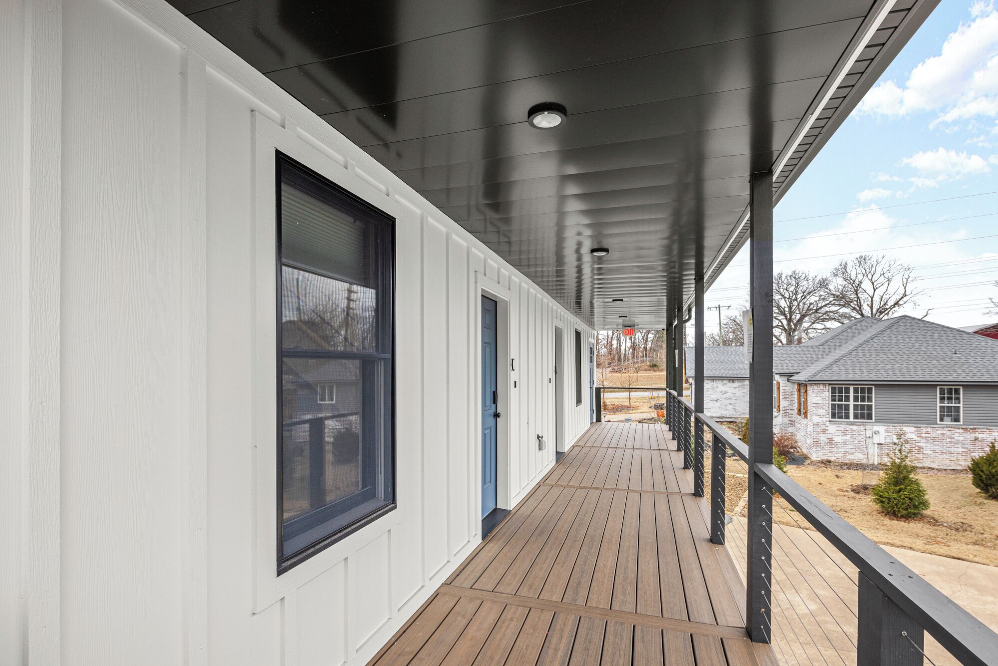 A covered porch with a wooden deck, a black railing, and a view of a residential area.