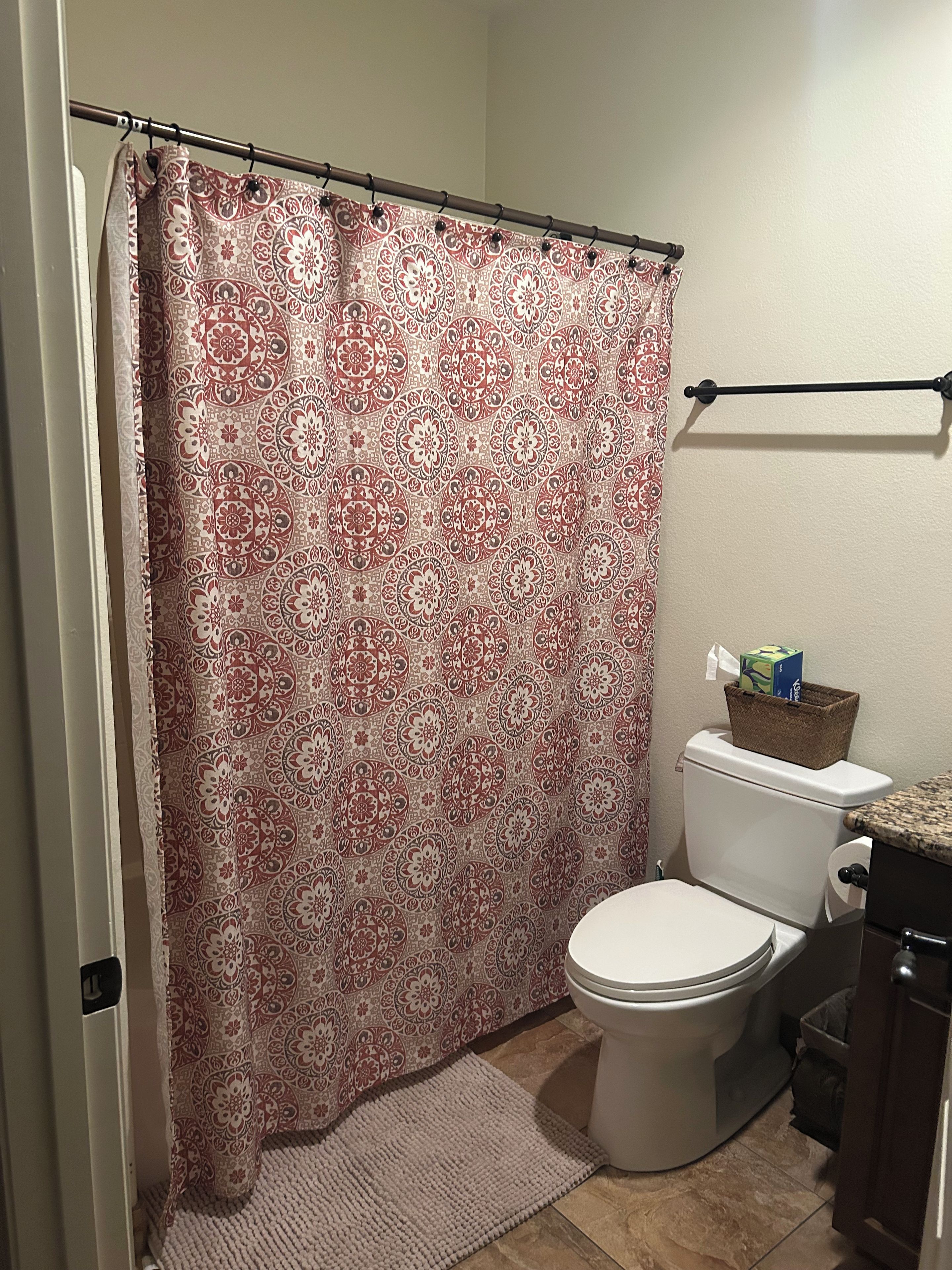 A bathroom with a patterned shower curtain, a toilet, a towel rack, and a small trash bin.
