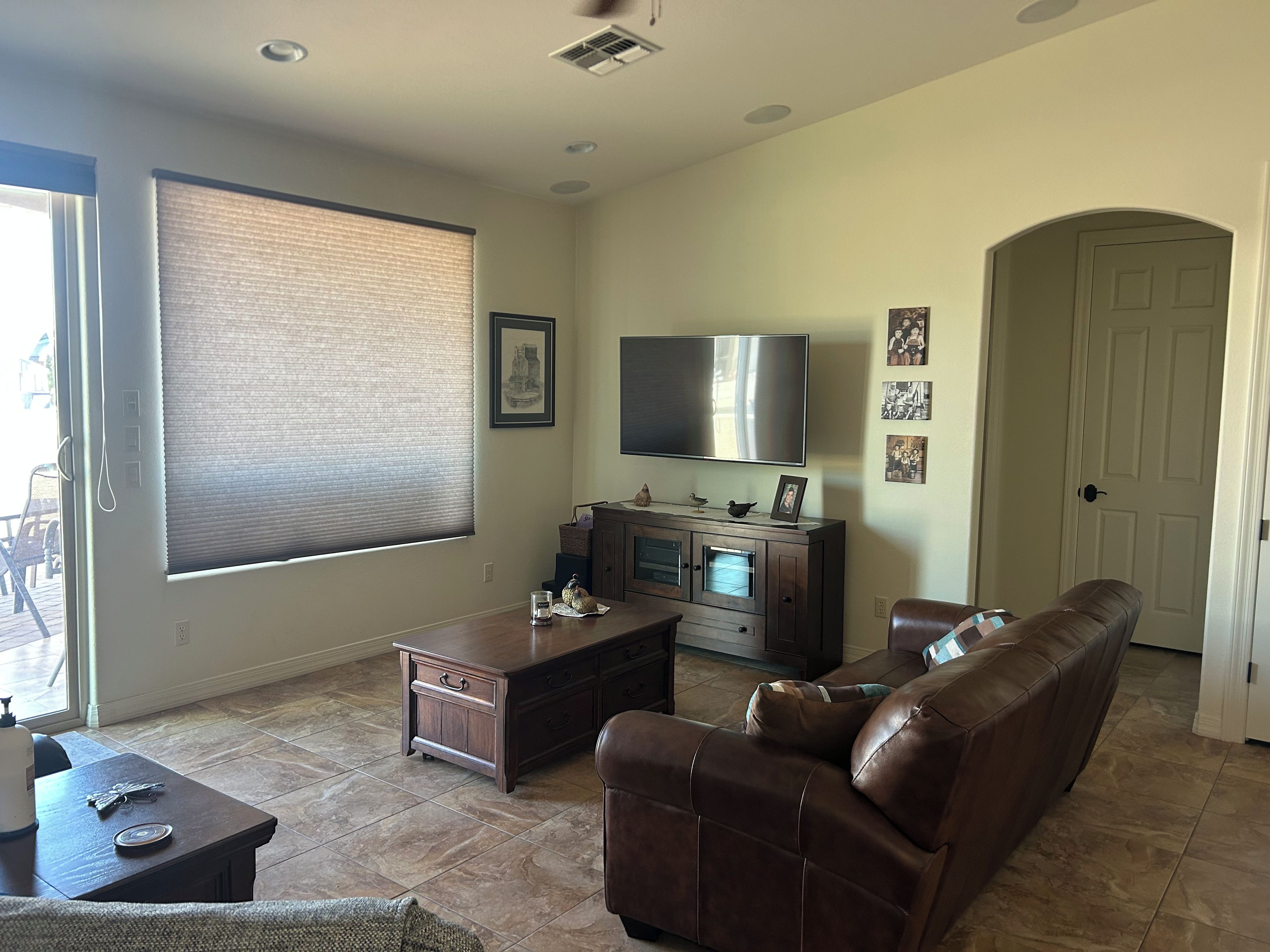 A living room with a brown leather sofa, a flat-screen TV, a wooden cabinet, and a large window with blinds.