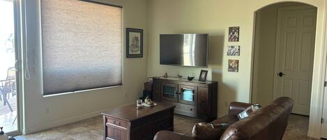 A living room with a brown leather sofa, a flat-screen TV, a wooden cabinet, and a large window with blinds.