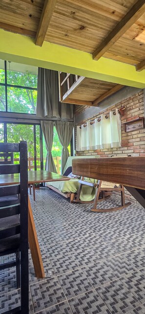 Ein Zimmer mit einem Bett, einer Holzbank und Blick auf Grün hinter großen Fenstern.