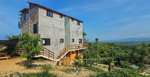 Edifici de diverses plantes amb una escala que condueix a un balcó, envoltat de vegetació i una vista de les colines llunyanes.
