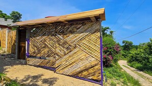 Cerca de bambu com uma porta, um caminho de terra e vegetação ao fundo.