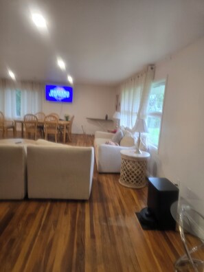 Een woonkamer met een televisie, houten vloeren en een eethoek met stoelen en een tafel.