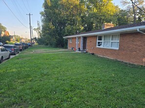 A residential area with a brick house, a grassy lawn, parked cars, and a row of trees. - Spacious Ranch 5-bedroom house with parking and garage on no outlet strret (Cincinnati)