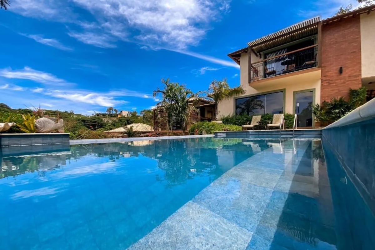 A swimming pool with clear blue water, surrounded by a landscaped area with palm trees and a modern house with a balcony.