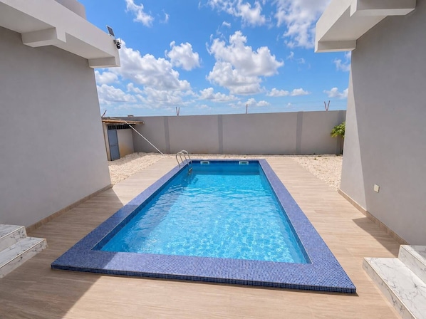 Piscina rectangular con borde de azulejos, rodeada por una terraza de tonos claros y un muro bajo.