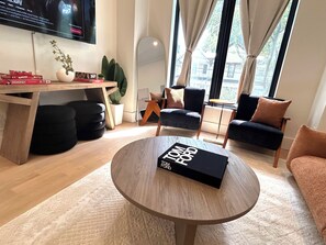 Un salon moderne avec une table basse ronde en bois, une bibliothèque, un téléviseur à écran plat fixé au mur et une vue sur l’extérieur grâce à de grandes fenêtres.