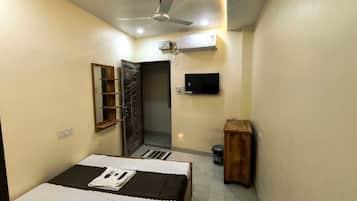 A compact bedroom with a bed, a ceiling fan, an air conditioning unit, a television, and a small wooden cabinet.