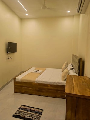 A bedroom with a bed, a wooden headboard, a bedside table, a wall-mounted TV, and a small rug. - OM BNB AC ROOMS Available in Varanasi  (Varanasi)