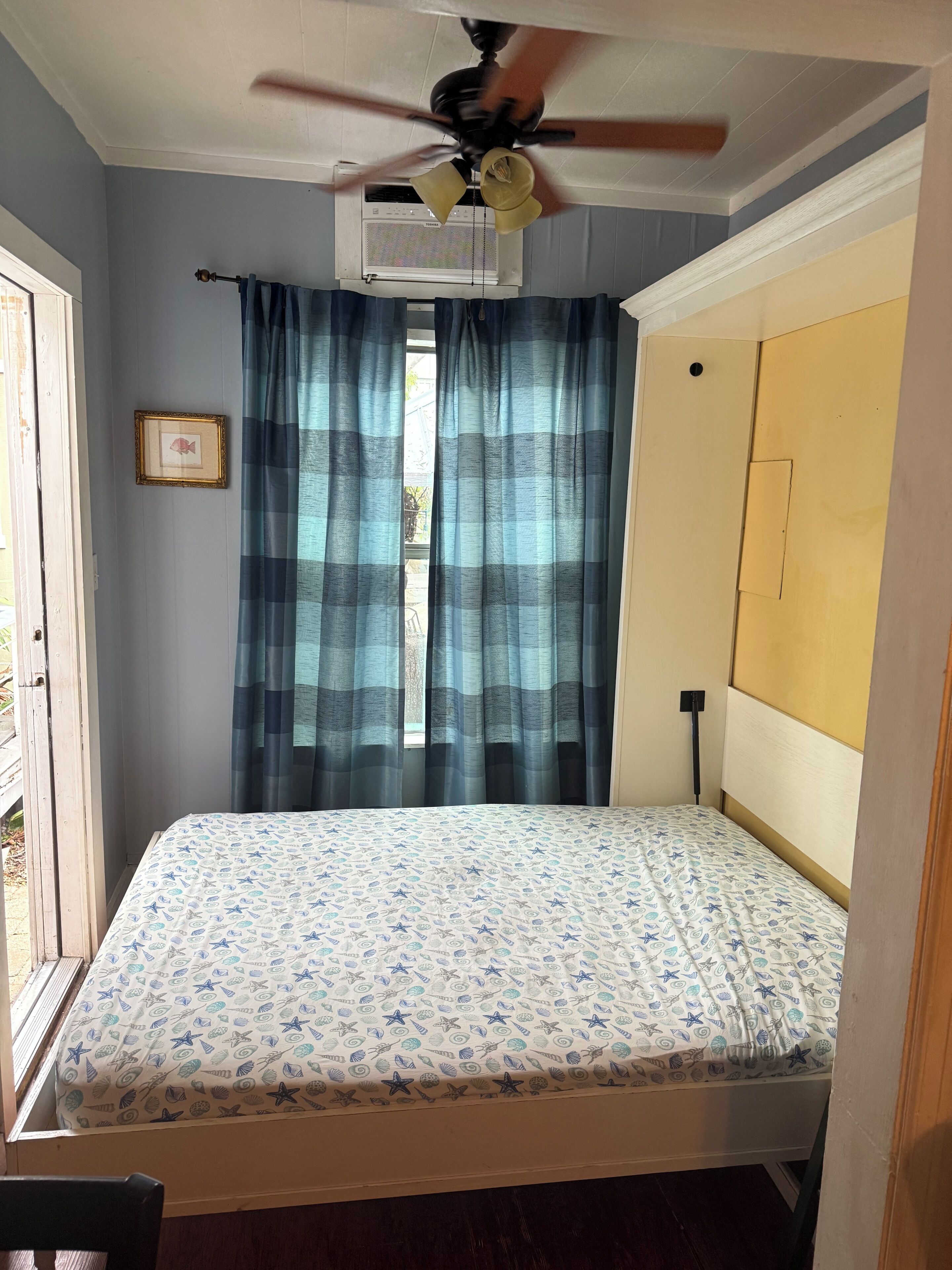 A bedroom with a bed, a ceiling fan, a window with curtains, and an air conditioning unit.