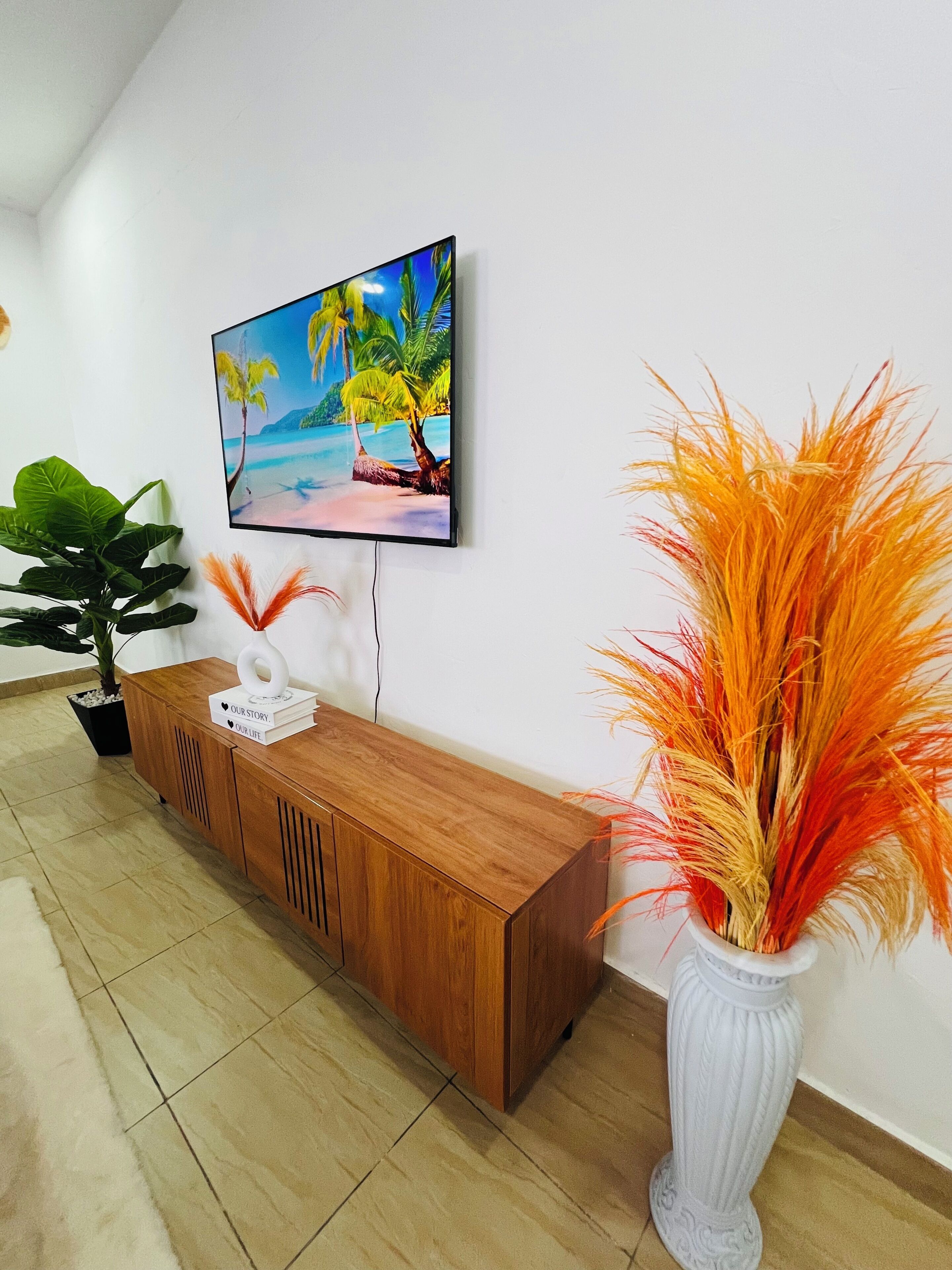 Un salon moderne avec un meuble TV en bois, un téléviseur fixé au mur qui diffuse une scène de plage, une plante en pot et un vase décoratif orné de feuillage orange.
