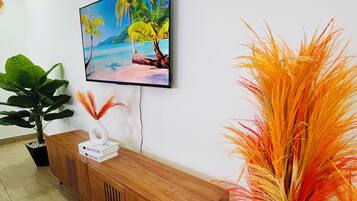 Un salon moderne avec un meuble TV en bois, un téléviseur fixé au mur qui diffuse une scène de plage, une plante en pot et un vase décoratif orné de feuillage orange.