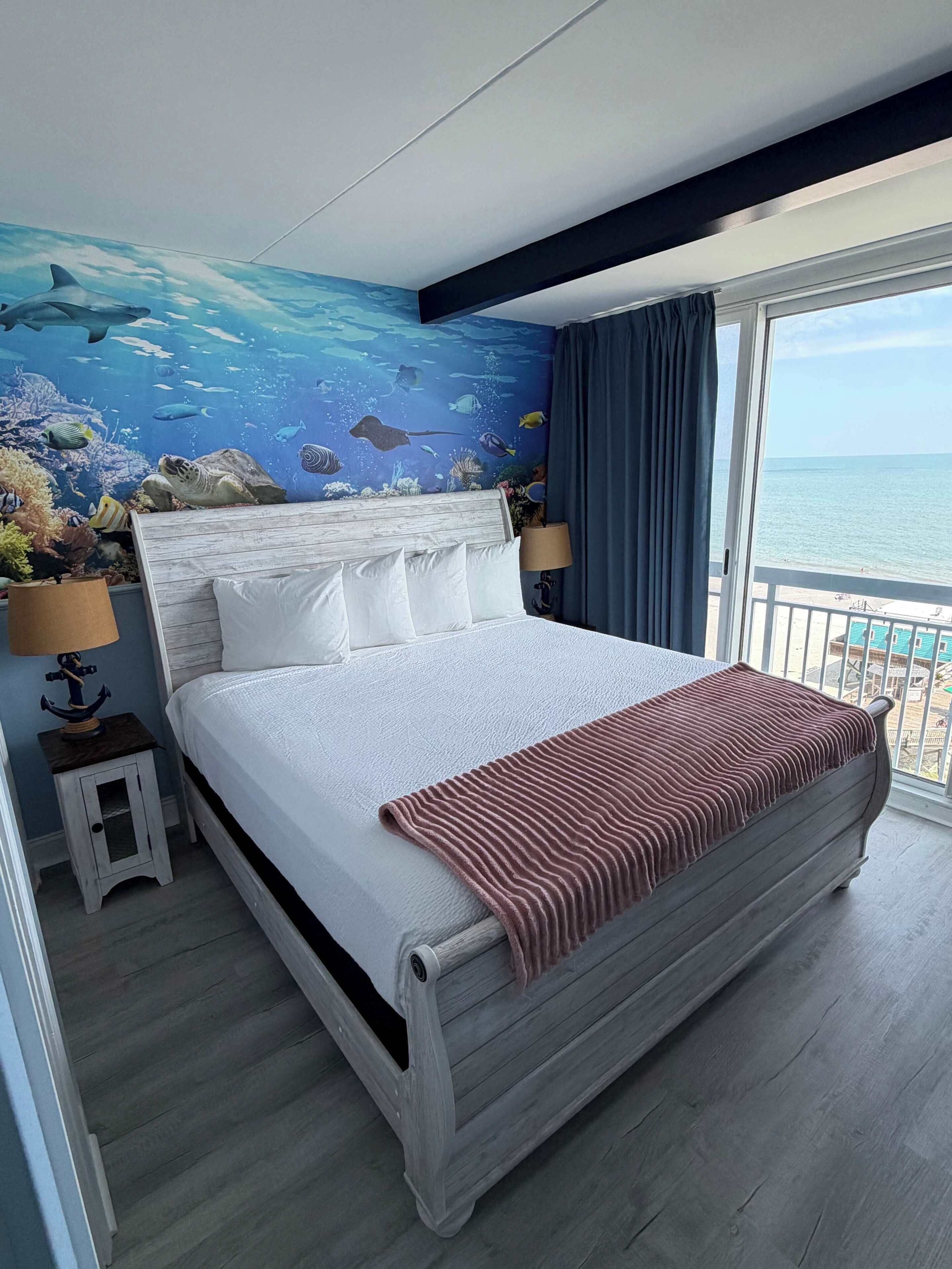 A bedroom with a bed, a nightstand, a lamp, a view of the sea through a window, and an ocean mural on the wall.
