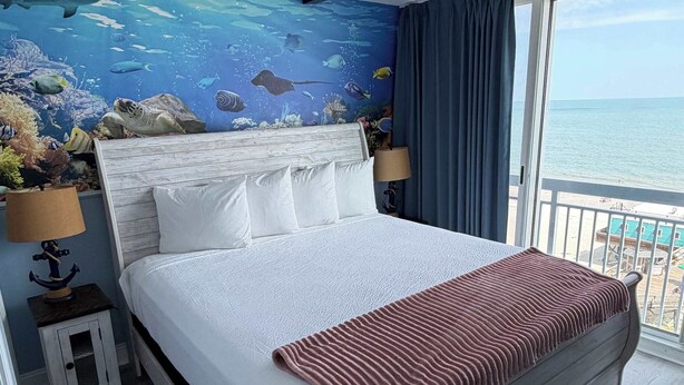 Ein Schlafzimmer mit einem Bett, einem Nachttisch, einer Lampe, Blick auf das Meer durch ein Fenster und einem Wandbild mit Meeresmotiv.