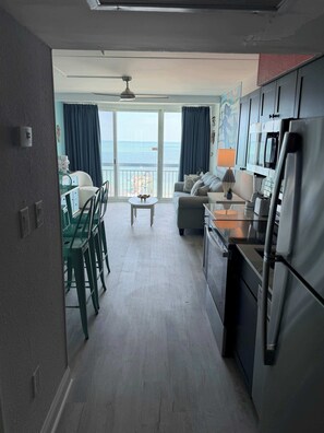 Eine moderne Küche mit Kühlschrank, Mikrowelle und Herd. Es gibt einen Essbereich mit Stühlen und Tisch sowie ein Wohnzimmer mit Sofa und Couchtisch. Ein Balkon mit Meerblick ist sichtbar.