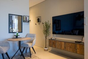 Ein modernes Wohnzimmer mit einem runden Holztisch, zwei grauen Sesseln, einem großen Flachbildfernseher an der Wand, einer Zierpflanze und einem schlichten Schrank.