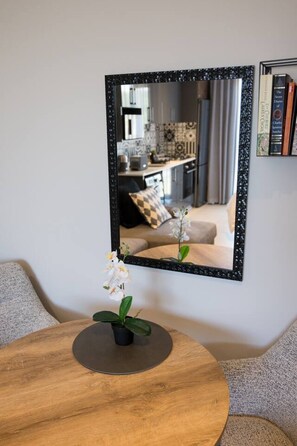 A round wooden table with a potted orchid, a mirror reflecting a living room with a sofa, a coffee table, and a kitchen area. - Slick Studio at Ocean Club H24 (Ballito)