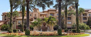 Un hotel de varios pisos con una fuente central, rodeado de palmeras y jardines cuidados.