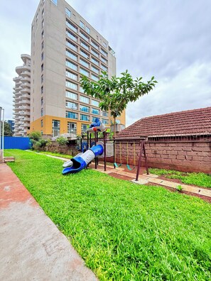 A playground with a slide and swings, surrounded by a grassy area and a brick wall. - Cozy 2-bedroom apartment with AC in fantastic Nairobi (Nairobi)