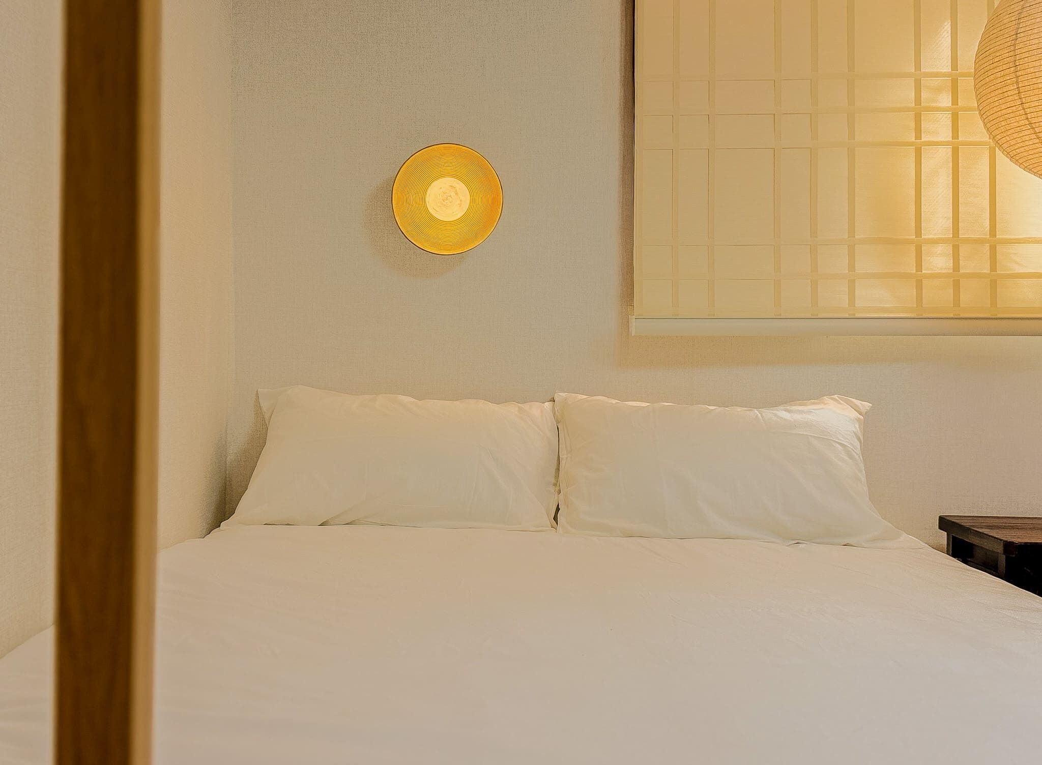 A bed with white bedding, a wall-mounted light fixture, and a window with blinds.