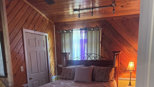 A bedroom with a wooden ceiling, a bed with a headboard, a nightstand, a lamp, and a window with curtains.