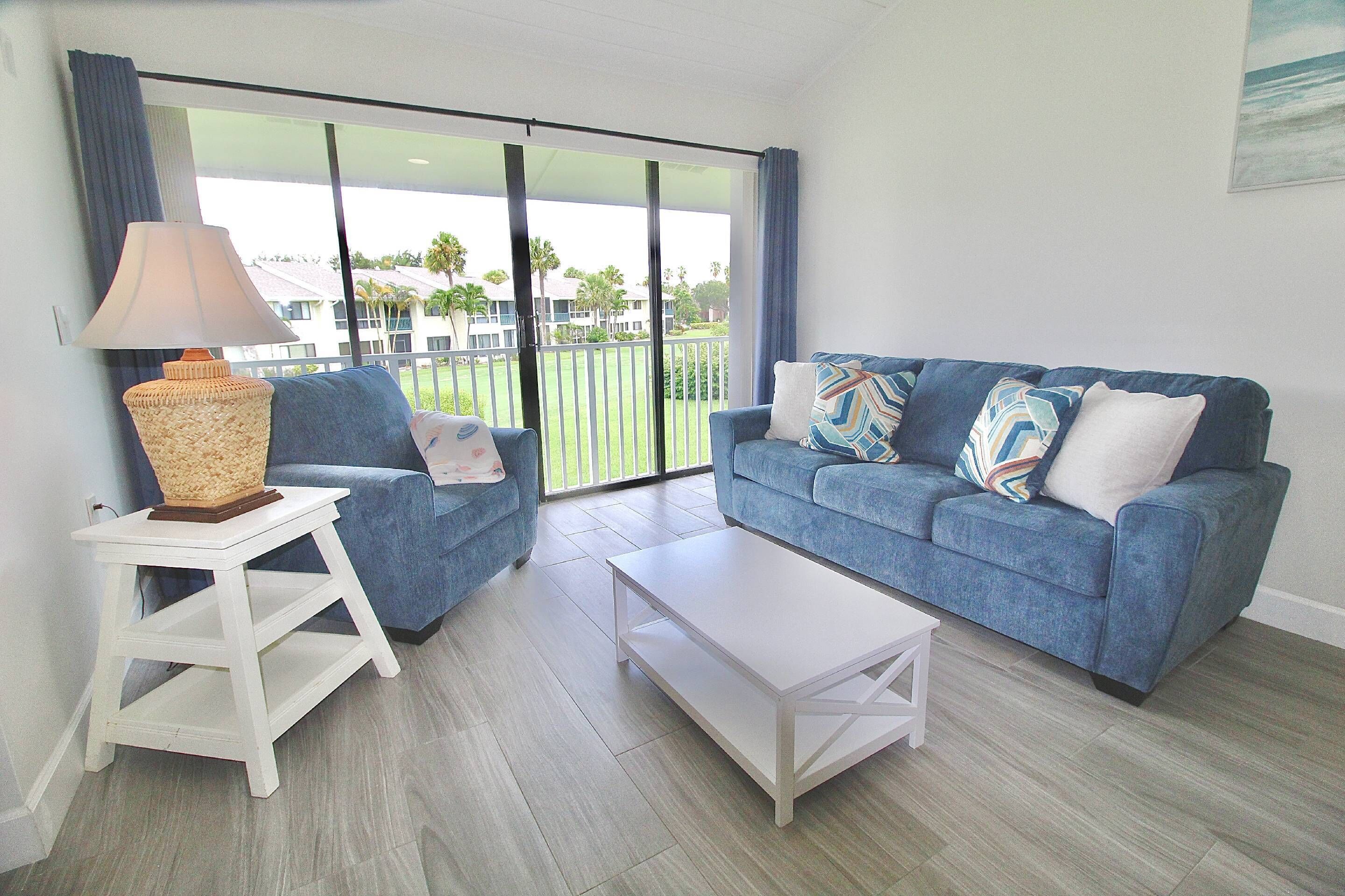 A living room with a blue sofa, a white coffee table, a lamp, and a view of a green landscape through large glass doors.