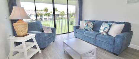 A living room with a blue sofa, a white coffee table, a lamp, and a view of a green landscape through large glass doors.