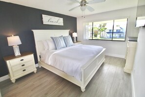 Une chambre à coucher comprenant un lit, des tables de chevet, un ventilateur de plafond, une fenêtre avec vue et un panneau fixé au mur.