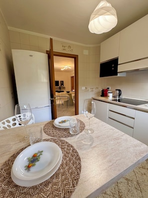 A modern kitchen with a dining area, featuring a refrigerator, a coffee maker, and a dining table set with plates and glasses.