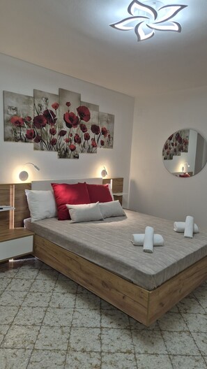 Une chambre moderne avec un lit surélevé en bois, des coussins rouges et blancs, un grand tableau floral, des lampes de chevet et un miroir rond.