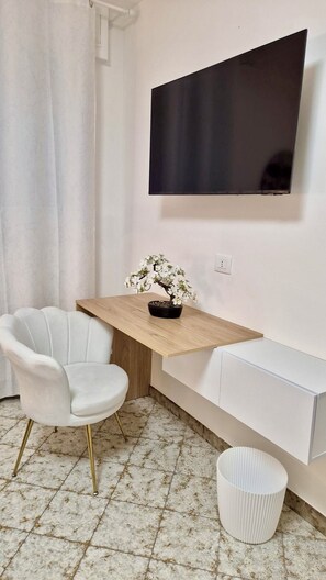 A small, modern room with a flat-screen TV mounted on the wall, a wooden desk with a potted plant, a white chair, and a radiator.