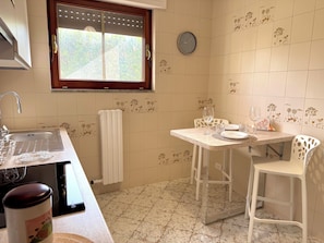 Une cuisine équipée d’un évier, d’une fenêtre, d’une horloge et d’un coin repas avec des chaises et une table.