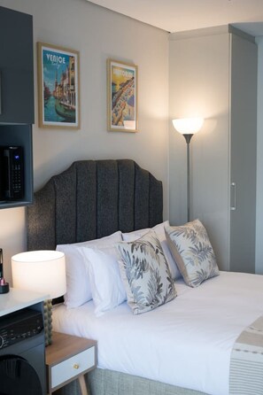 Quarto de hotel com uma cama, uma escrivaninha, um abajur e duas molduras na parede.
