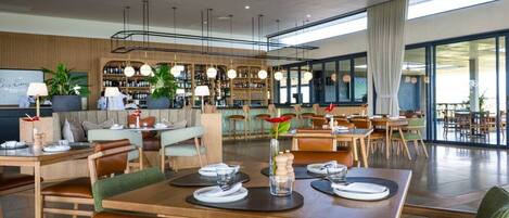 A modern restaurant with wooden tables and chairs, a bar area, and large windows allowing natural light.