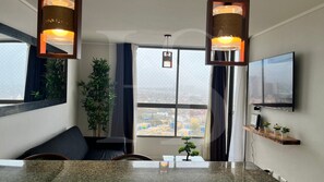 A modern hotel room with a large window, a flat-screen TV, a wooden shelf with plants, and a small table.