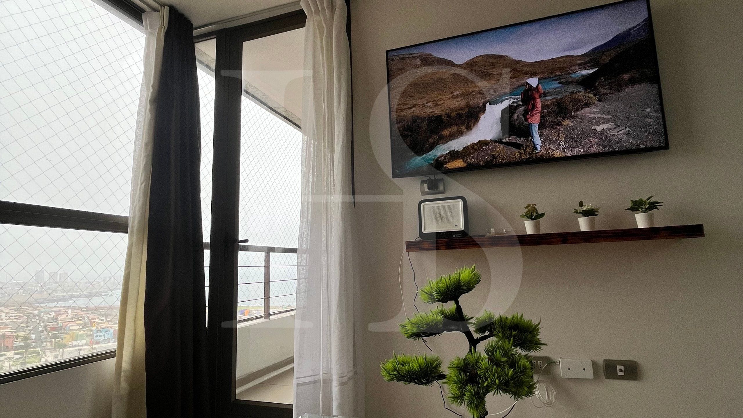 Una habitación con una televisión montada en la pared, una planta pequeña en un estante y una ventana con cortinas.