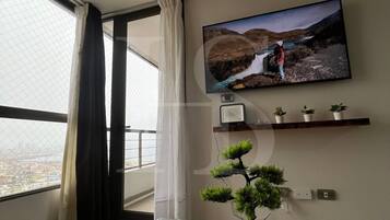Una habitación con una televisión montada en la pared, una planta pequeña en un estante y una ventana con cortinas.