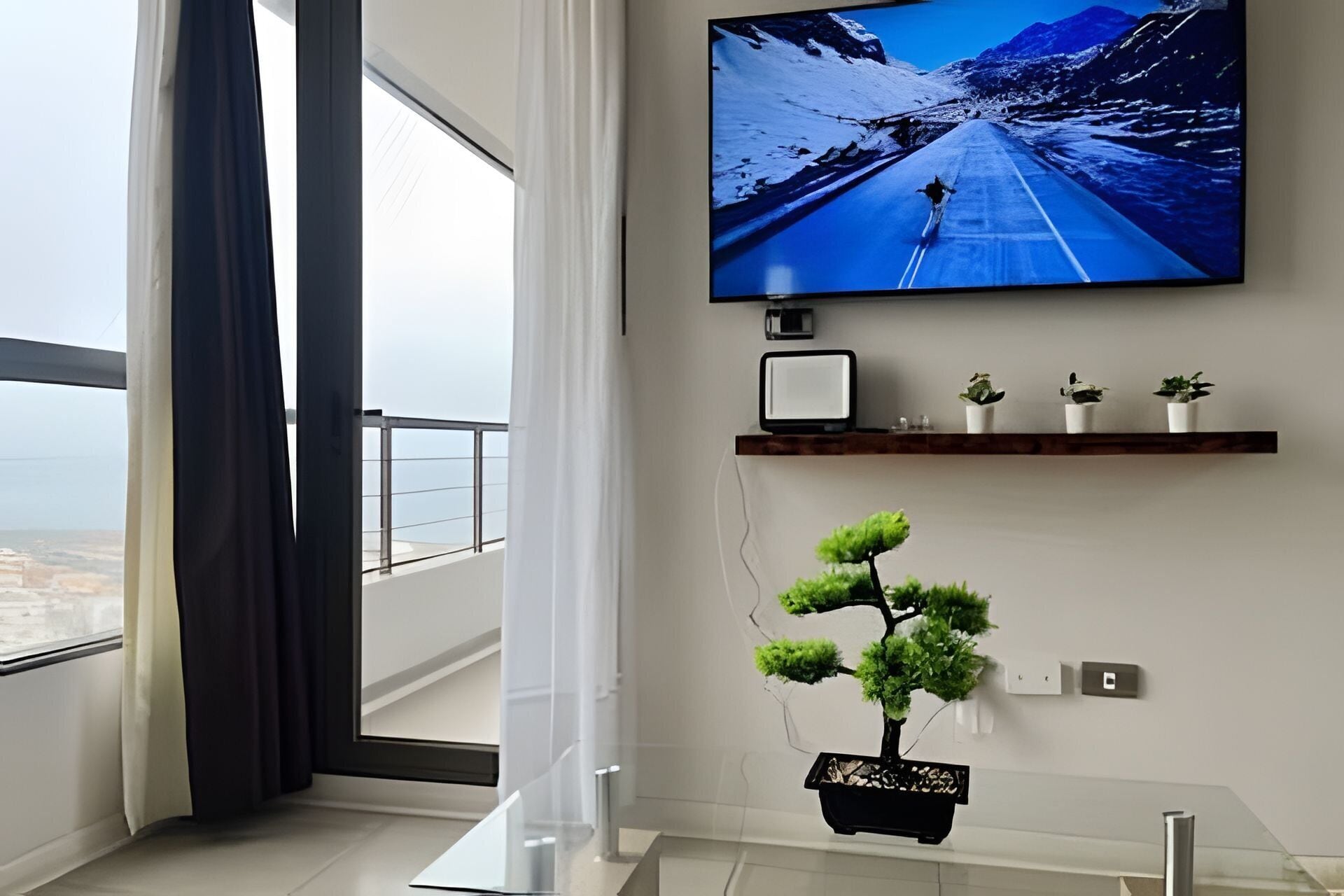 Una sala de estar moderna con un ventanal grande, un televisor de pantalla plana montado en la pared, un estante con objetos decorativos y un bonsái.