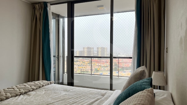 A bedroom with a bed, a nightstand, a lamp, and a view of buildings through a window with curtains.