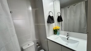 Un baño con lavamanos blanco, inodoro, ducha y espejo con barra para toallas.