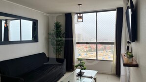 A living room with a black sofa, a glass coffee table, a potted plant, a hanging light fixture, and a large window with curtains.
