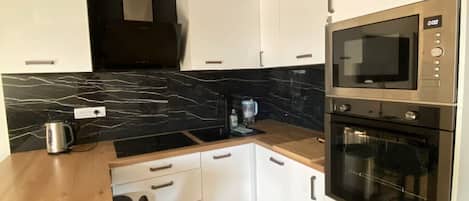 Cocina compacta con armarios blancos, encimera negra y electrodomésticos de acero inoxidable, incluyendo un microondas y un horno.